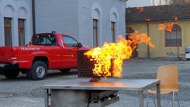 Brennendes Modell eines PC-Bildschirms auf einem Tisch im Freien. Im Hintergrund steht ein Feuerwehrauto.